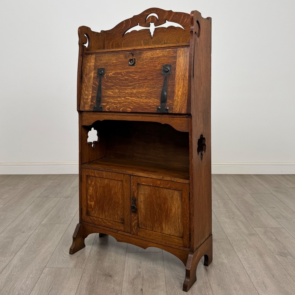 An antique Arts and Crafts oak writing bureau featuring a slant drop-front desk, a pierced cresting top rail, dark iron strap hardware, an open shelf, and lower double doors. View IV