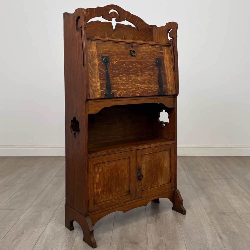 An antique Arts and Crafts oak writing bureau featuring a slant drop-front desk, a pierced cresting top rail, dark iron strap hardware, an open shelf, and lower double doors. View X