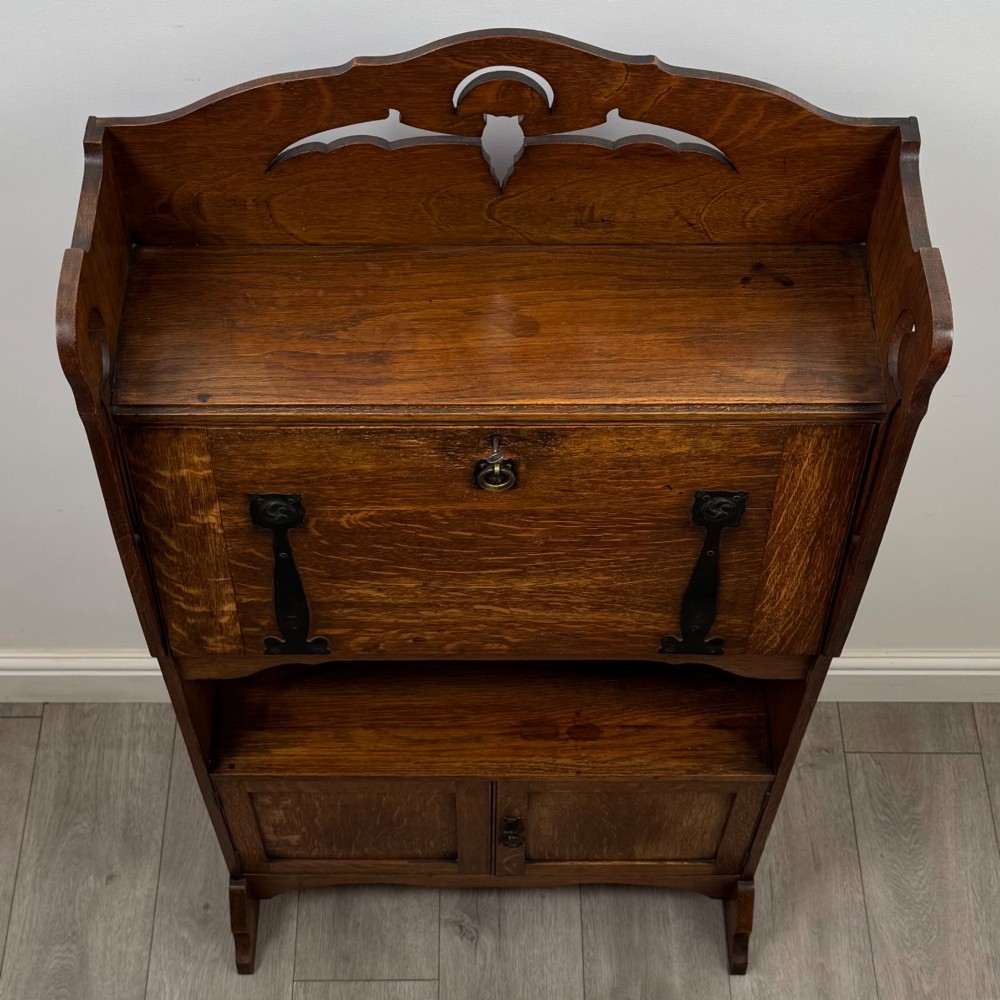 An antique Arts and Crafts oak writing bureau featuring a slant drop-front desk, a pierced cresting top rail, dark iron strap hardware, an open shelf, and lower double doors. View XI