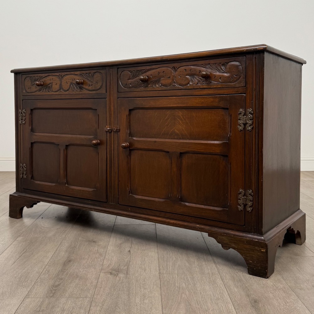 A dark oak sideboard featuring two carved upper drawers above panelled cupboard doors with decorative iron hinges and wooden knobs. View III