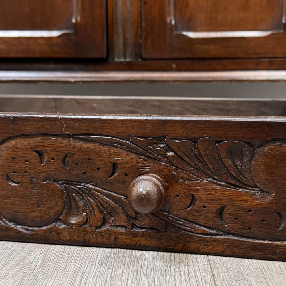 A dark oak sideboard featuring two carved upper drawers above panelled cupboard doors with decorative iron hinges and wooden knobs. View XII