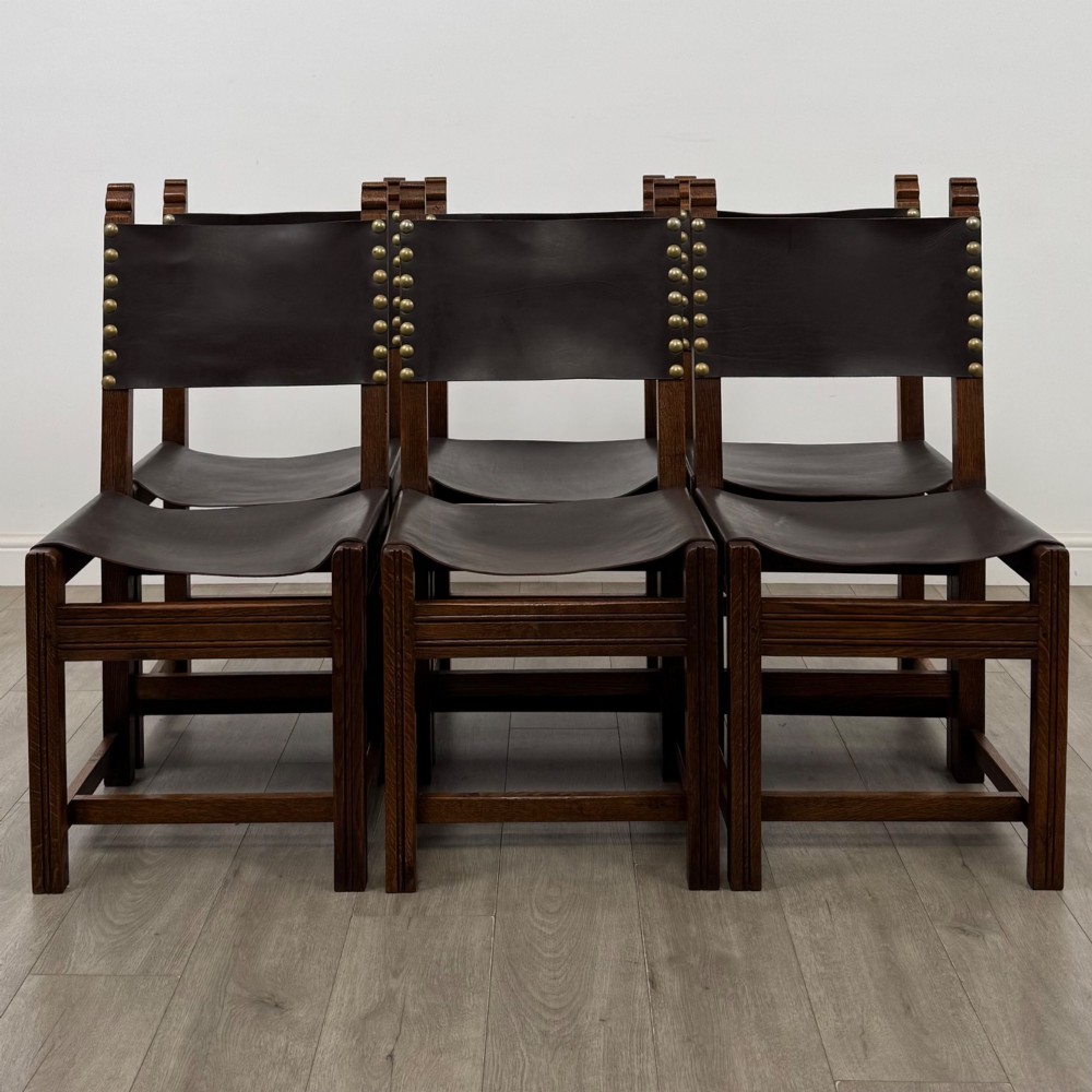 A set of six dark oak dining chairs featuring thick dark brown leather sling backrests and seats secured with decorative brass studs.