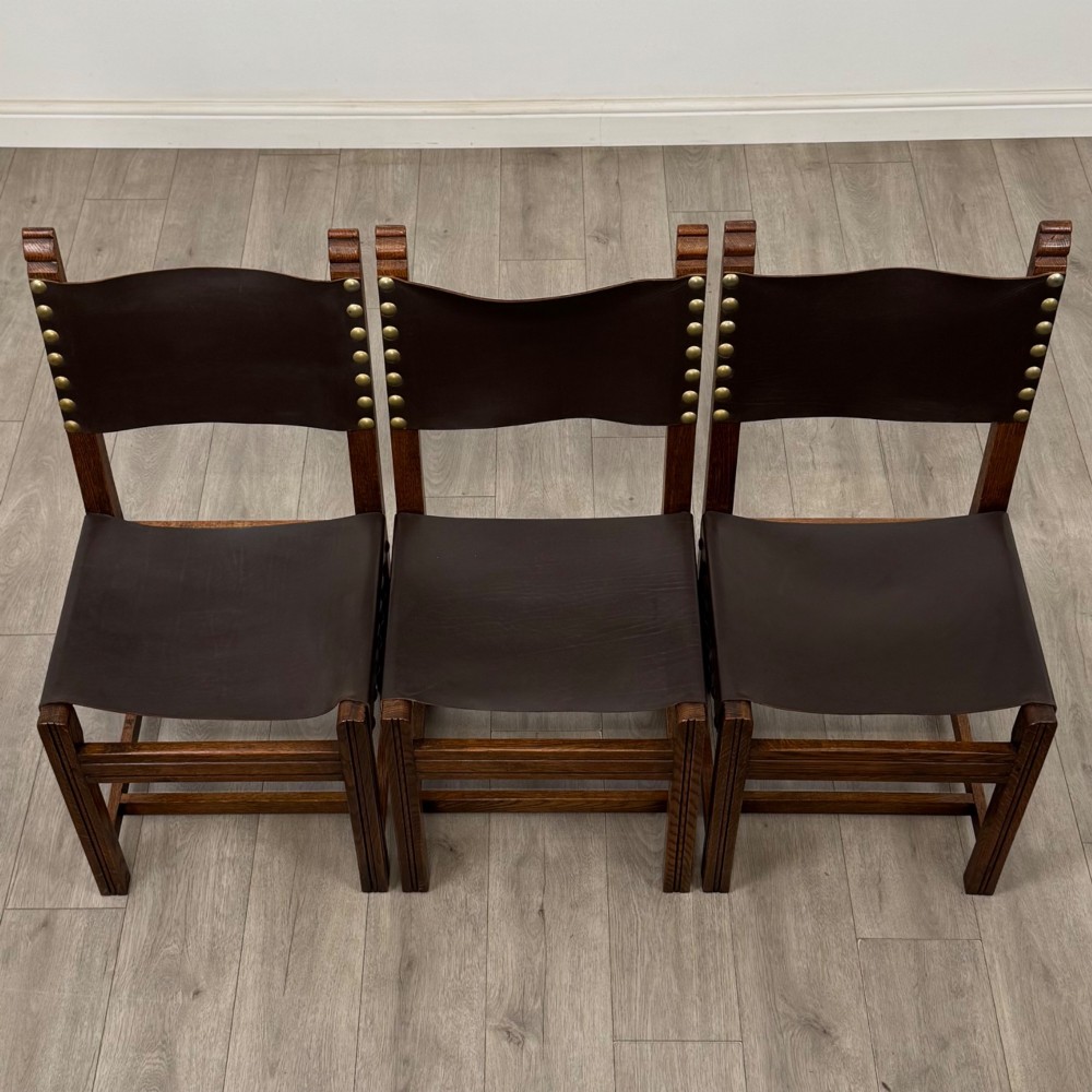 A set of six dark oak dining chairs featuring thick dark brown leather sling backrests and seats secured with decorative brass studs. View IX