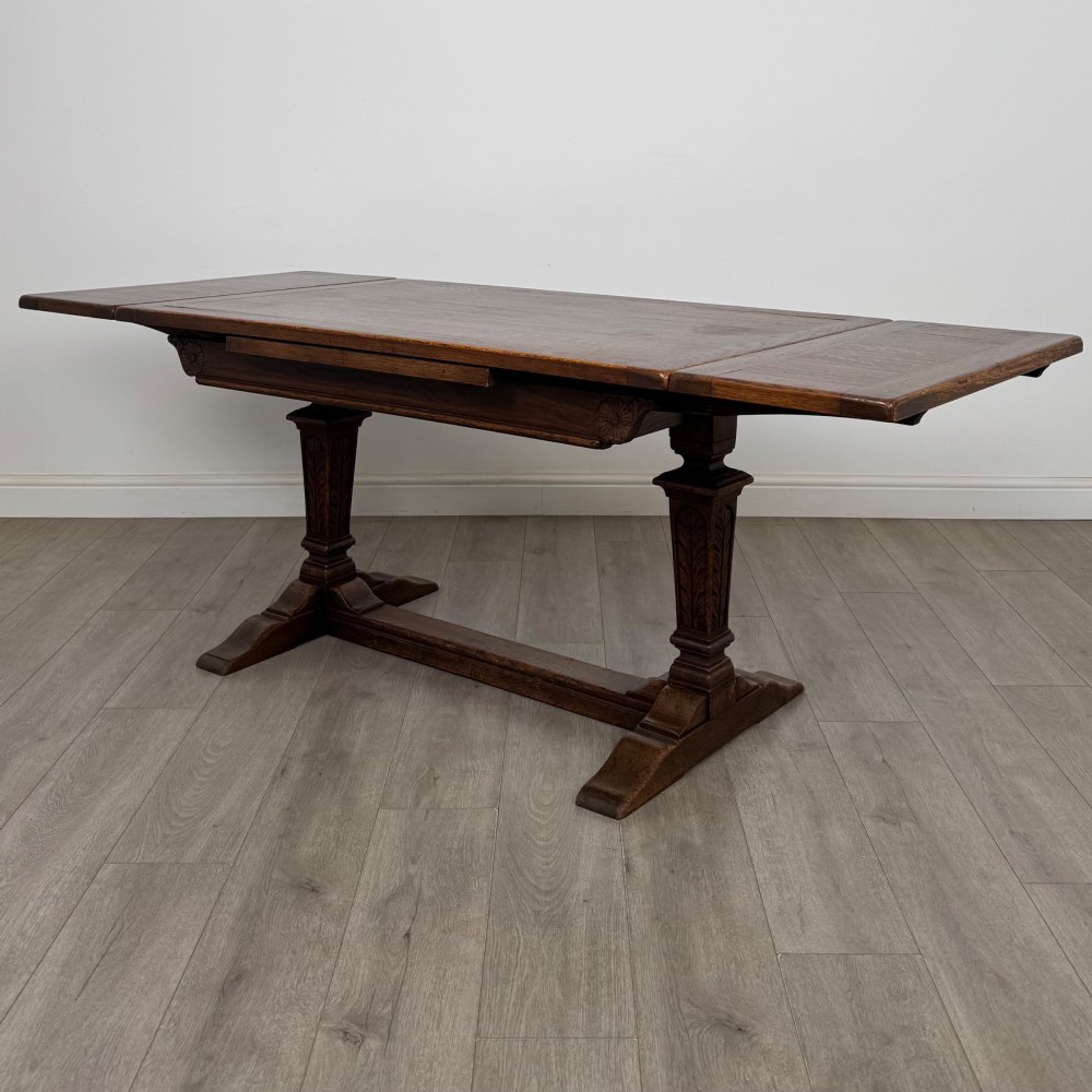 A dark oak draw leaf dining table featuring two extendable leaves, carved pedestal supports, and a low connecting stretcher base.