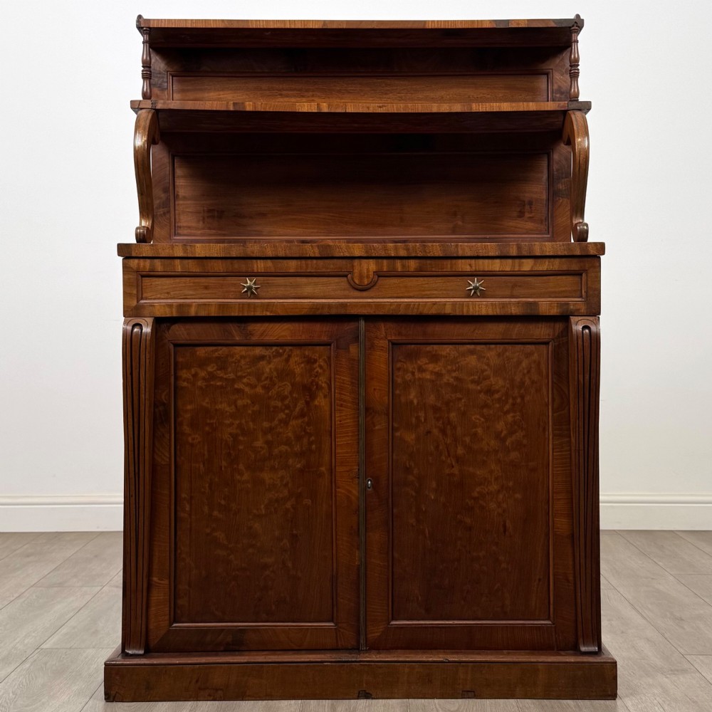 An antique Regency mahogany chiffonier featuring an upper two-tier shelf structure, a long drawer with star-shaped brass pulls, and a lower panelled double cupboard on a plinth base.