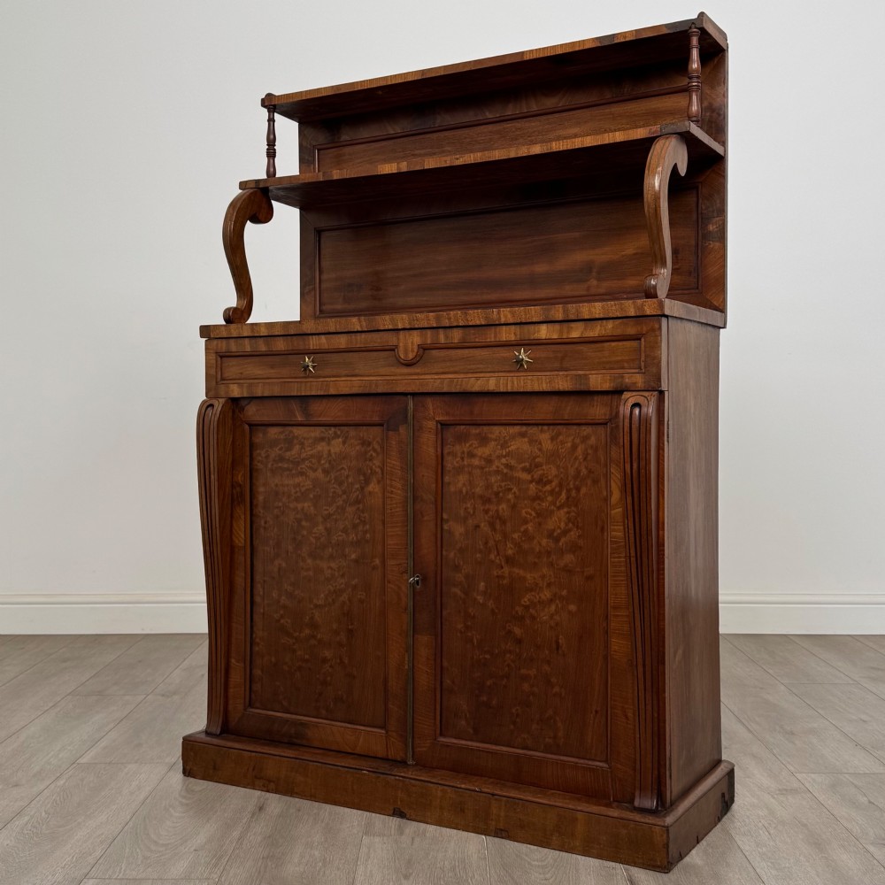 An antique Regency mahogany chiffonier featuring an upper two-tier shelf structure, a long drawer with star-shaped brass pulls, and a lower panelled double cupboard on a plinth base. View III