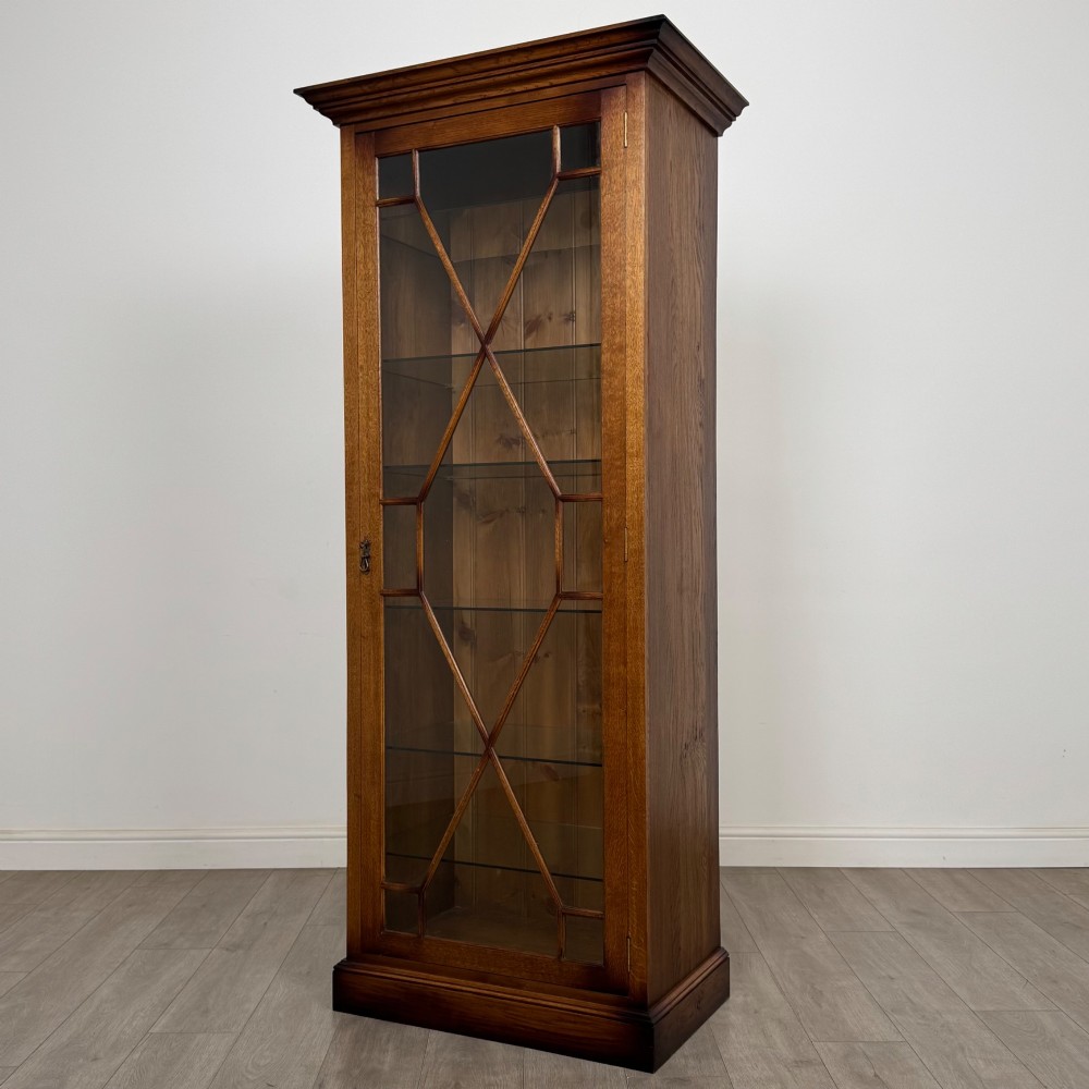 A tall 20th-century warm brown oak display cabinet featuring a single glass door with diamond-pattern astragal glazing bars, a moulded top cornice, and a solid plinth base. View IV