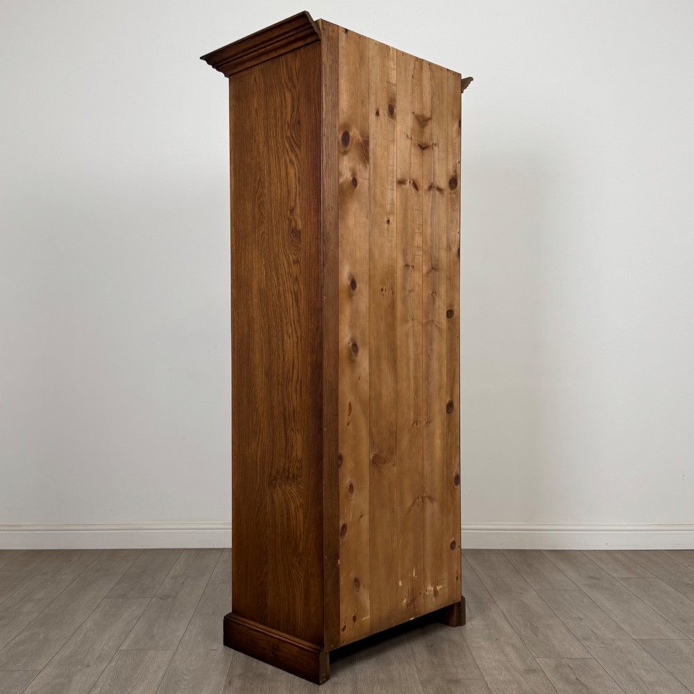 A tall 20th-century warm brown oak display cabinet featuring a single glass door with diamond-pattern astragal glazing bars, a moulded top cornice, and a solid plinth base. View VI
