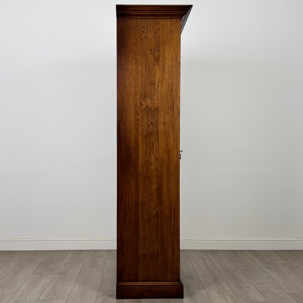 A tall 20th-century warm brown oak display cabinet featuring a single glass door with diamond-pattern astragal glazing bars, a moulded top cornice, and a solid plinth base. View IX