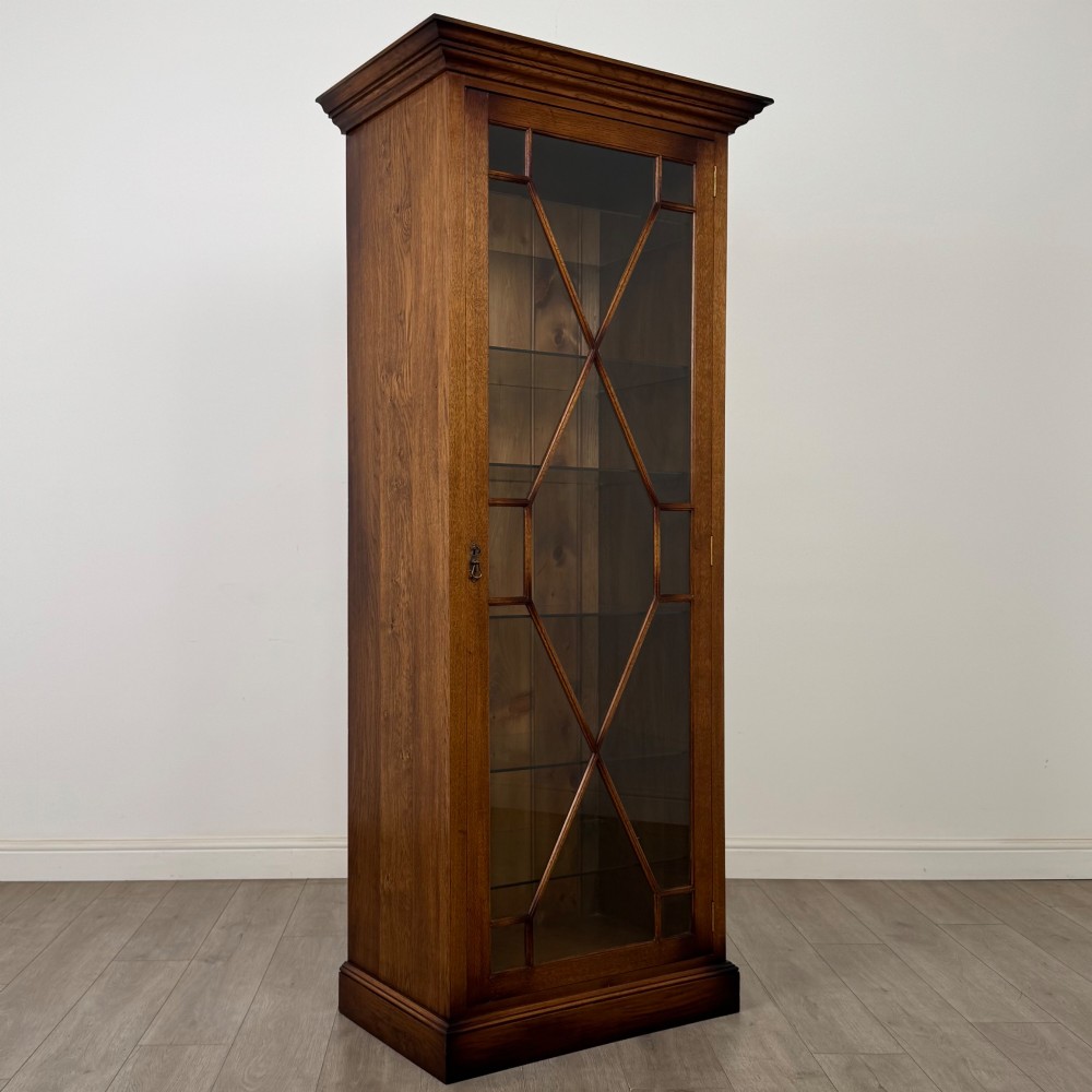 A tall 20th-century warm brown oak display cabinet featuring a single glass door with diamond-pattern astragal glazing bars, a moulded top cornice, and a solid plinth base. View X