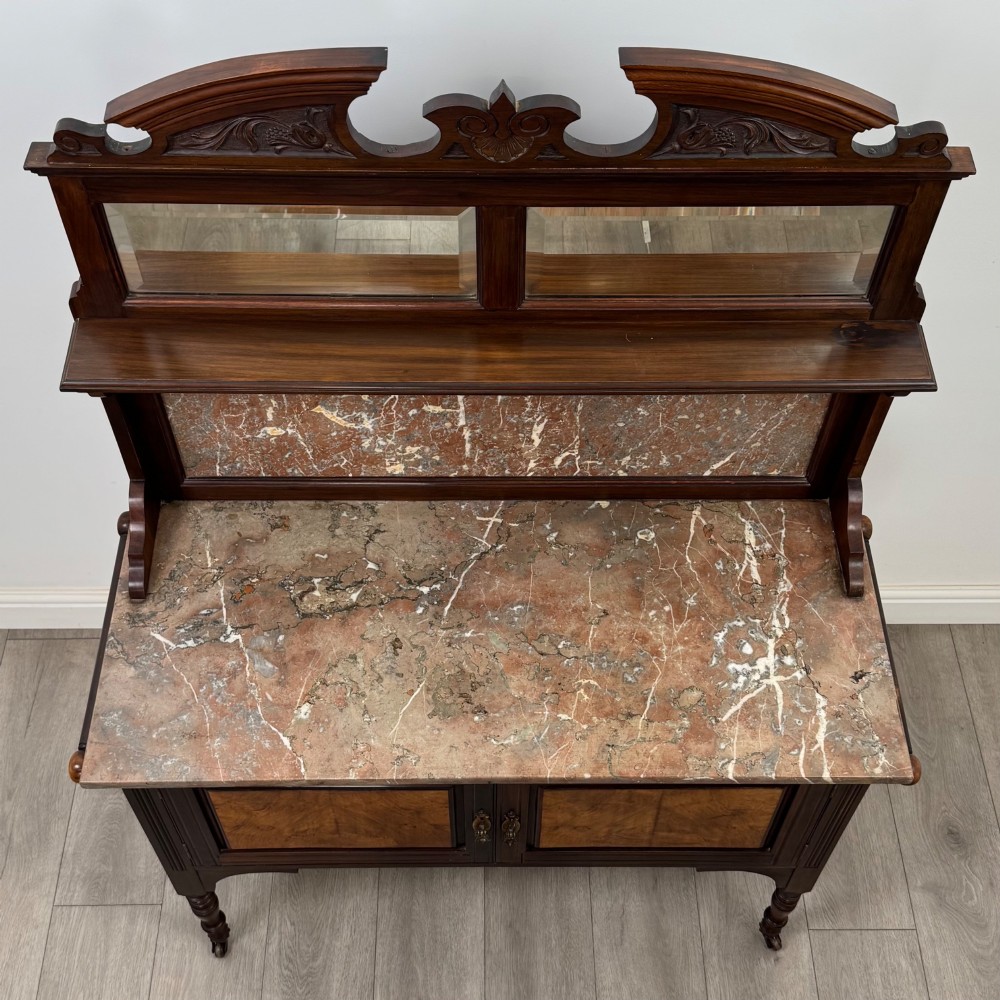 An Edwardian walnut washstand featuring a red marble splashback, mirror gallery top, double cupboard doors with burr walnut panels, and turned legs on castors. View X