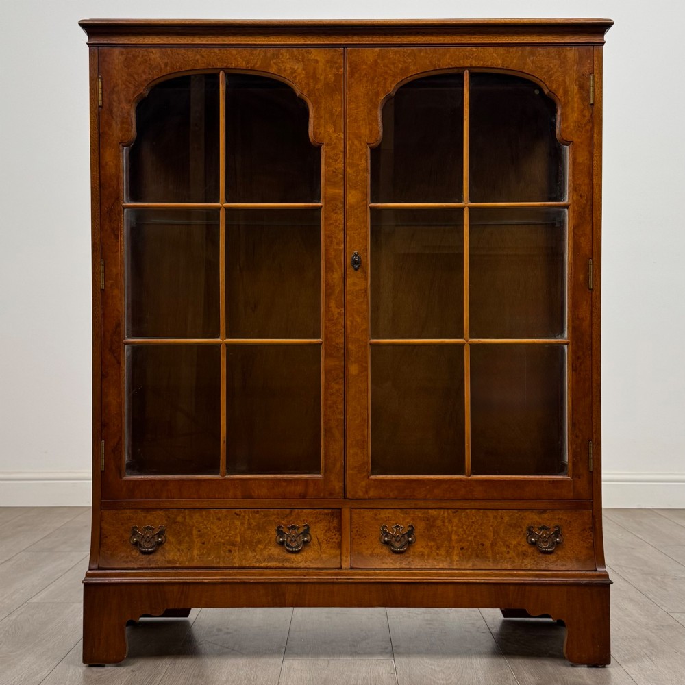 A 20th-century burr walnut display cabinet featuring two glazed doors with arched detail above two drawers on bracket feet.