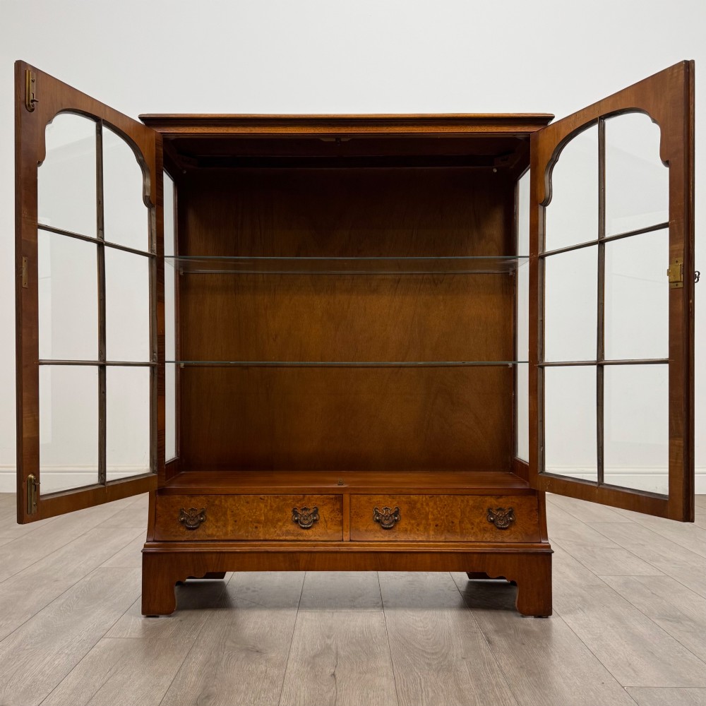 A 20th-century burr walnut display cabinet featuring two glazed doors with arched detail above two drawers on bracket feet. View II
