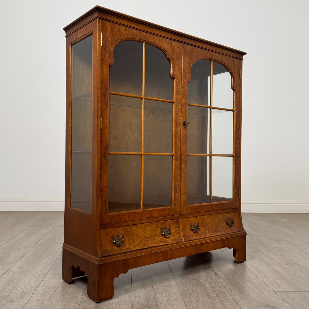 A 20th-century burr walnut display cabinet featuring two glazed doors with arched detail above two drawers on bracket feet. View IX