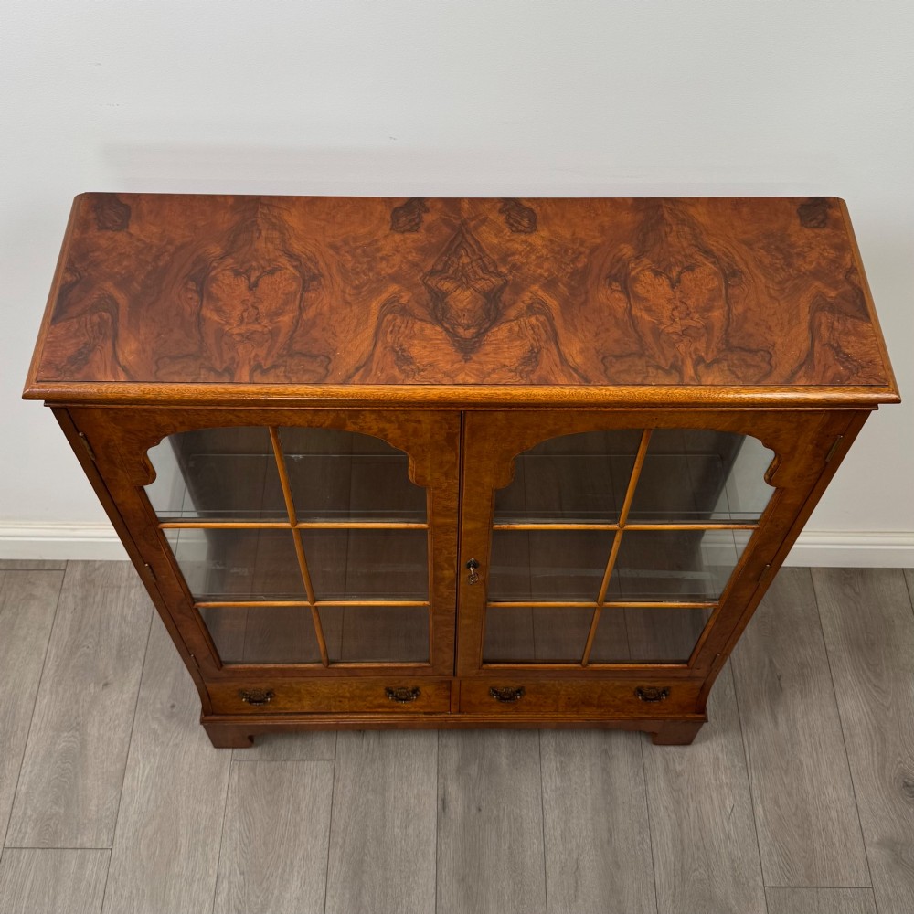 A 20th-century burr walnut display cabinet featuring two glazed doors with arched detail above two drawers on bracket feet. View X