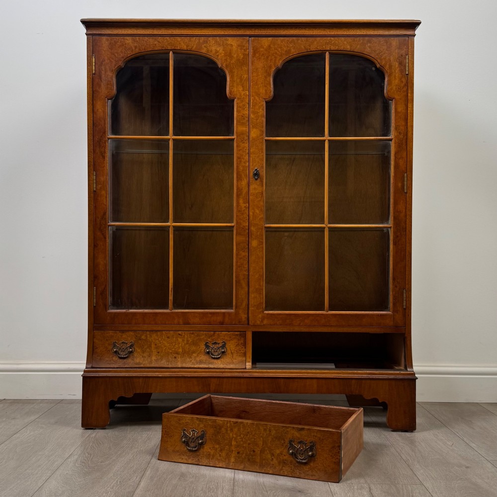 A 20th-century burr walnut display cabinet featuring two glazed doors with arched detail above two drawers on bracket feet. View XI