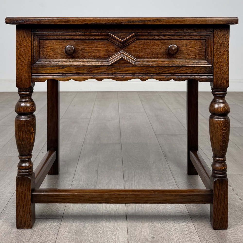 A dark oak side table featuring a single drawer with geometric mitred moulding, turned wooden knobs, a shaped lower apron, and baluster turned legs linked by low stretchers.