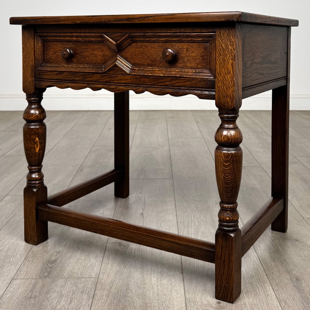 A dark oak side table featuring a single drawer with geometric mitred moulding, turned wooden knobs, a shaped lower apron, and baluster turned legs linked by low stretchers. View II