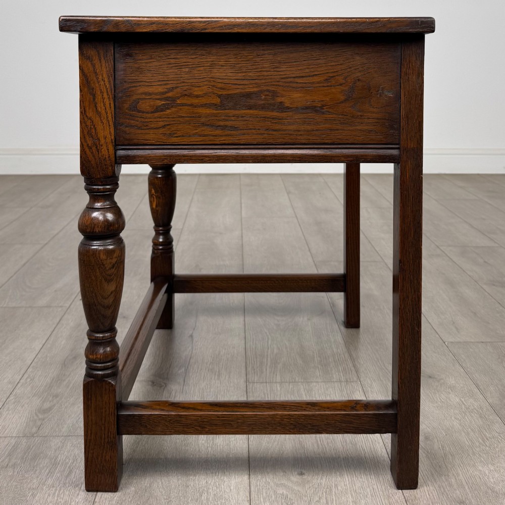 A dark oak side table featuring a single drawer with geometric mitred moulding, turned wooden knobs, a shaped lower apron, and baluster turned legs linked by low stretchers. View III