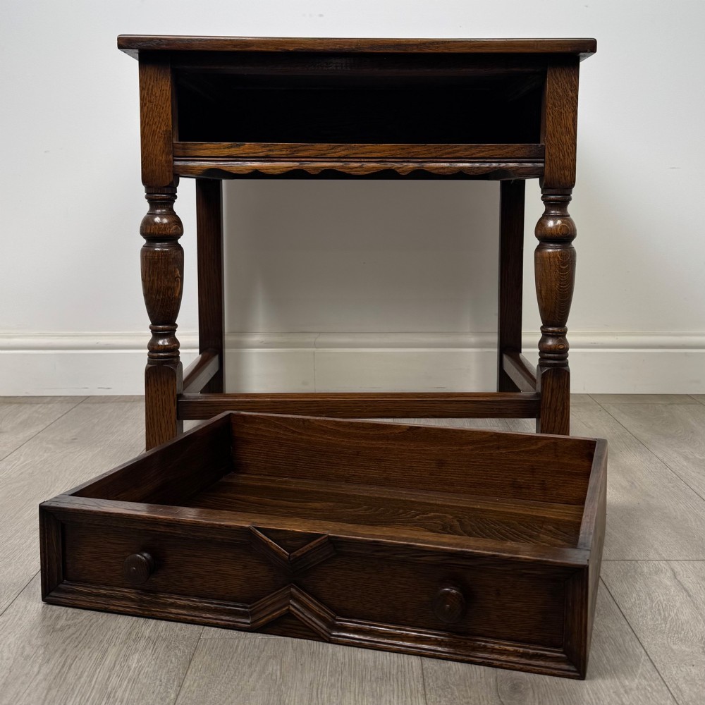 A dark oak side table featuring a single drawer with geometric mitred moulding, turned wooden knobs, a shaped lower apron, and baluster turned legs linked by low stretchers. View X