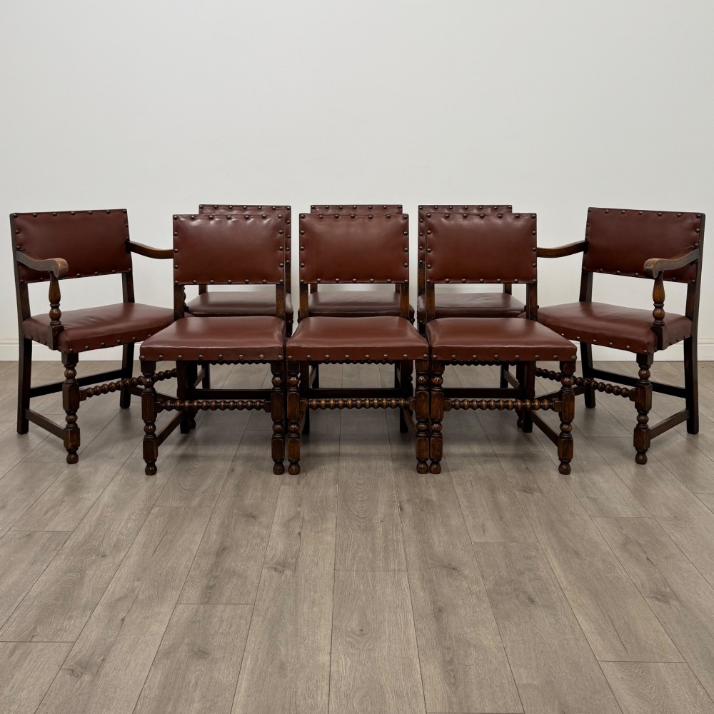 A set of eight dark oak dining chairs featuring brown leather upholstery secured with brass studding, turned legs, and including two armchairs.
