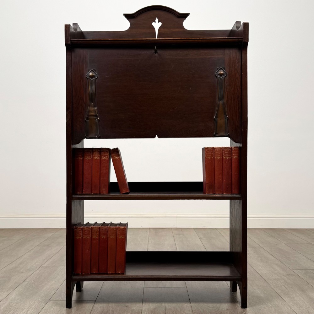 A dark mahogany Art Nouveau style bureau featuring a drop-front writing flap with elongated metal strap hinges above two open shelves.