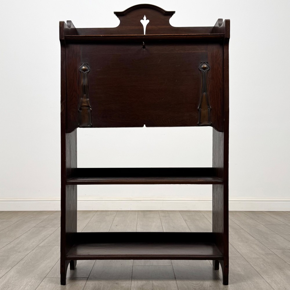 A dark mahogany Art Nouveau style bureau featuring a drop-front writing flap with elongated metal strap hinges above two open shelves. View II