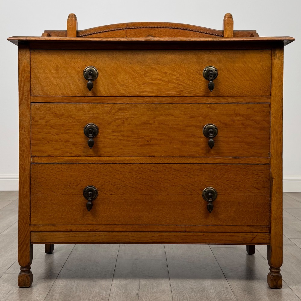 An antique Edwardian oak chest featuring three full-width drawers with dark metal drop handles and a shaped gallery back with turned finials.