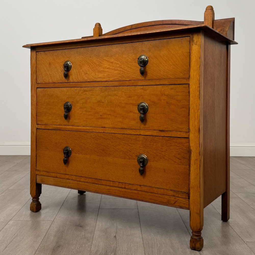 An antique Edwardian oak chest featuring three full-width drawers with dark metal drop handles and a shaped gallery back with turned finials. View II