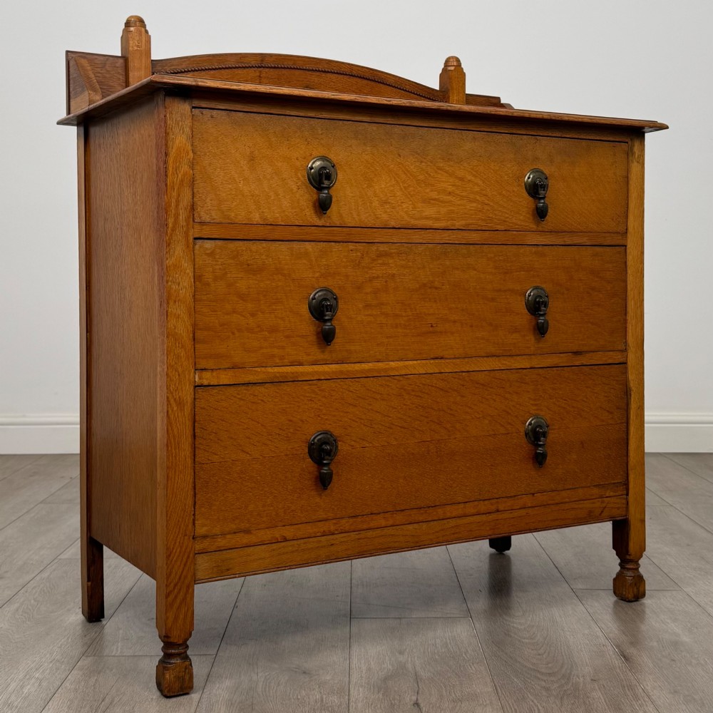 An antique Edwardian oak chest featuring three full-width drawers with dark metal drop handles and a shaped gallery back with turned finials. View VIII