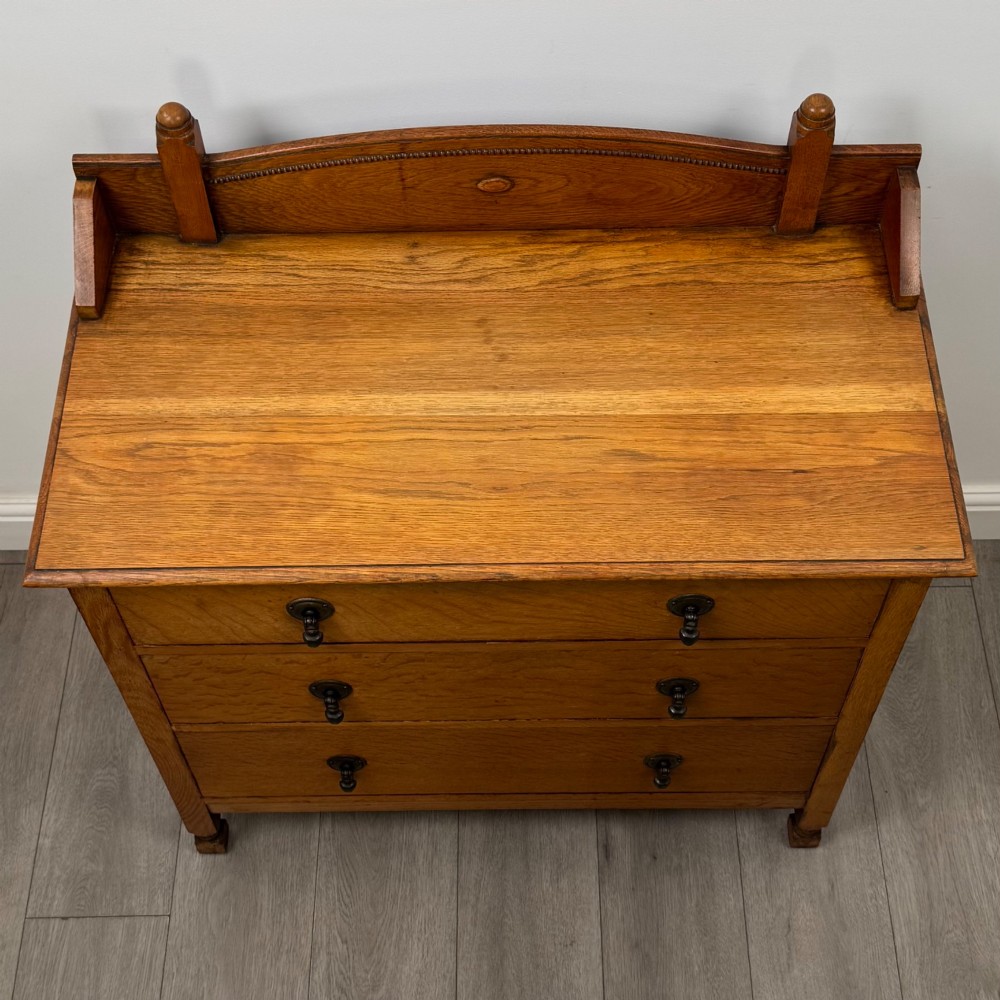 An antique Edwardian oak chest featuring three full-width drawers with dark metal drop handles and a shaped gallery back with turned finials. View IX