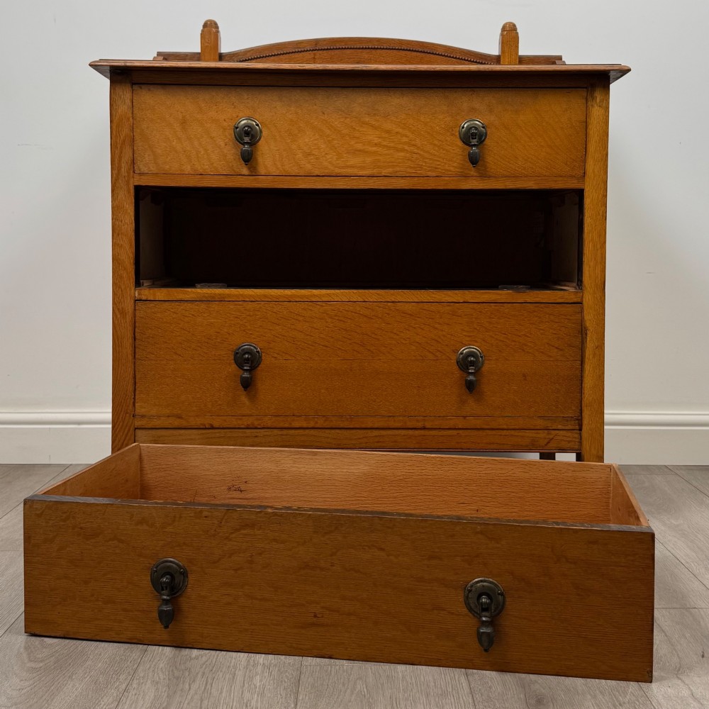 An antique Edwardian oak chest featuring three full-width drawers with dark metal drop handles and a shaped gallery back with turned finials. View X