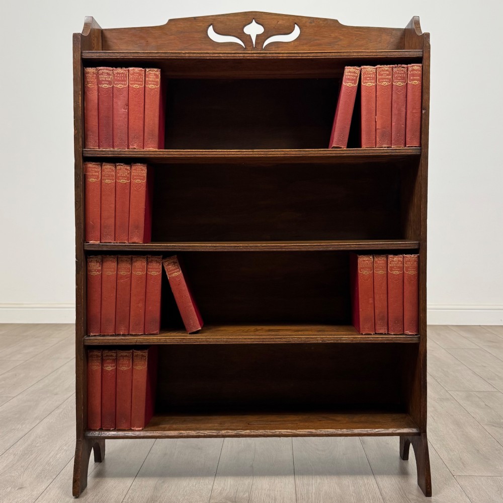 An early 20th-century Arts and Crafts medium oak open bookcase featuring four fixed shelves and a pierced stylised heart motif cut into the arched gallery top.