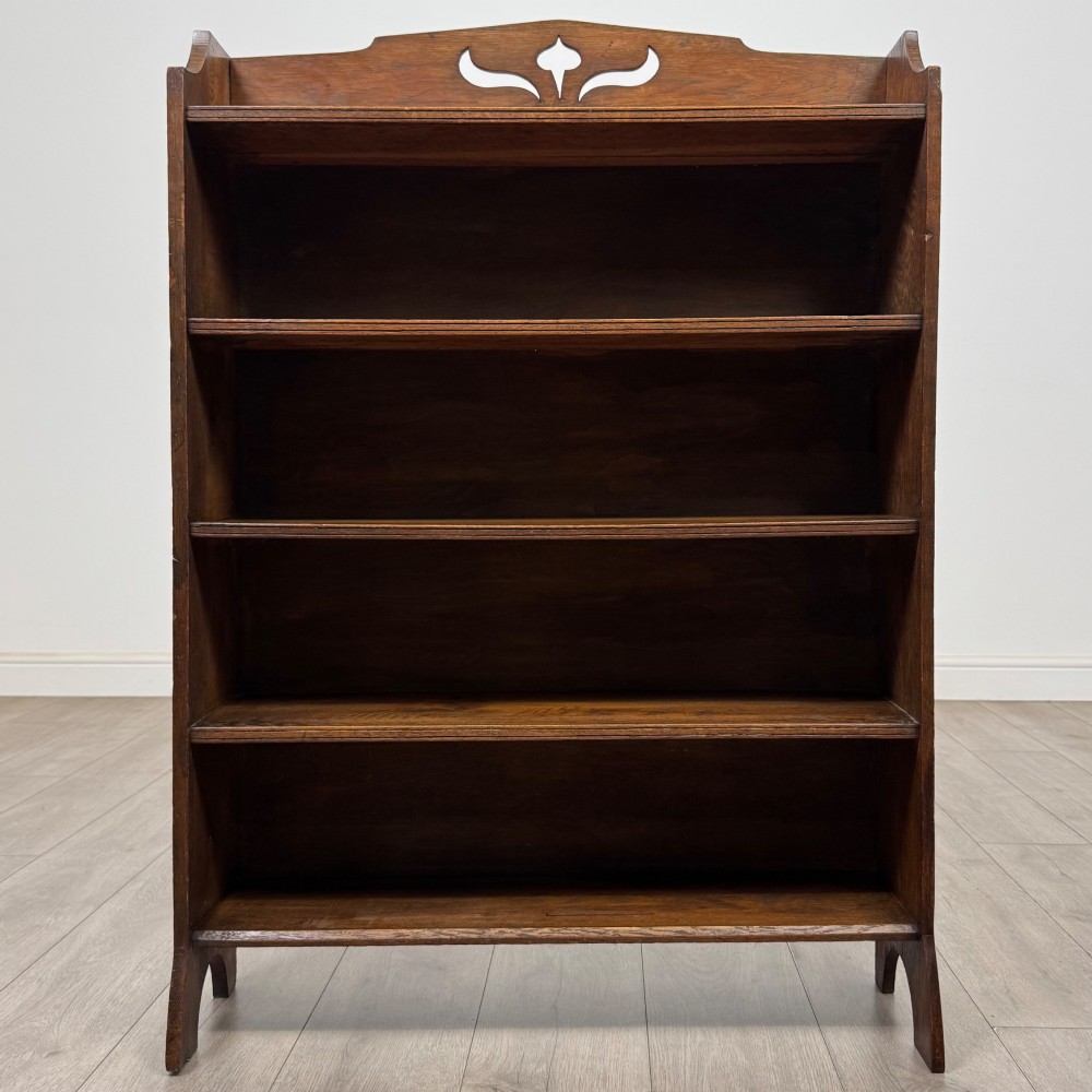 An early 20th-century Arts and Crafts medium oak open bookcase featuring four fixed shelves and a pierced stylised heart motif cut into the arched gallery top. View II