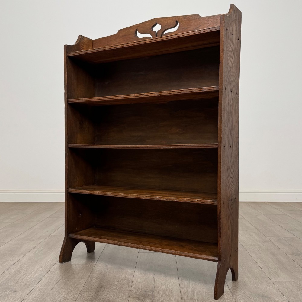 An early 20th-century Arts and Crafts medium oak open bookcase featuring four fixed shelves and a pierced stylised heart motif cut into the arched gallery top. View III