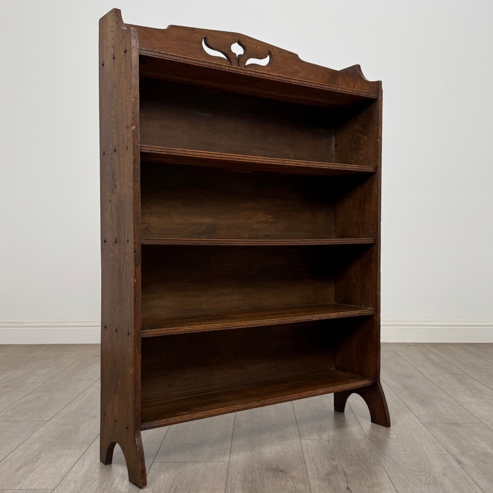 An early 20th-century Arts and Crafts medium oak open bookcase featuring four fixed shelves and a pierced stylised heart motif cut into the arched gallery top. View IX