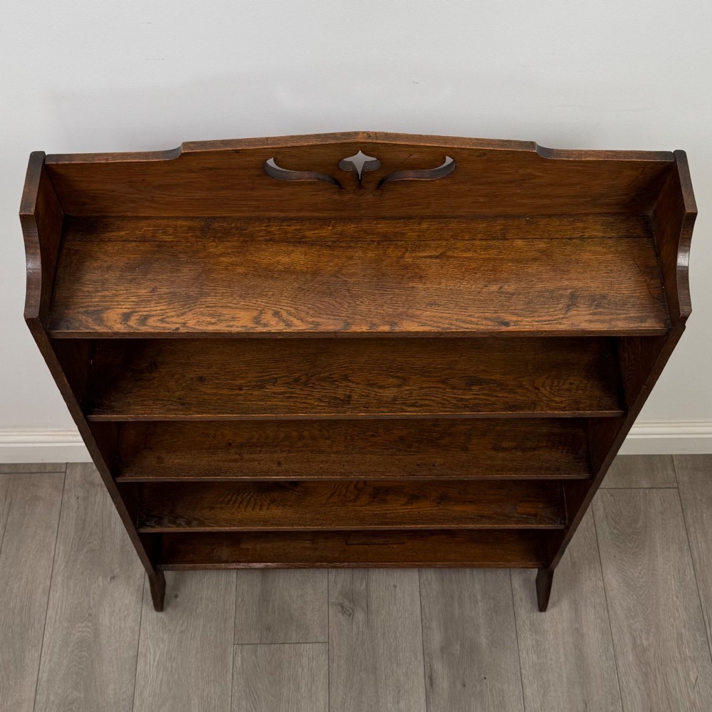 An early 20th-century Arts and Crafts medium oak open bookcase featuring four fixed shelves and a pierced stylised heart motif cut into the arched gallery top. View X
