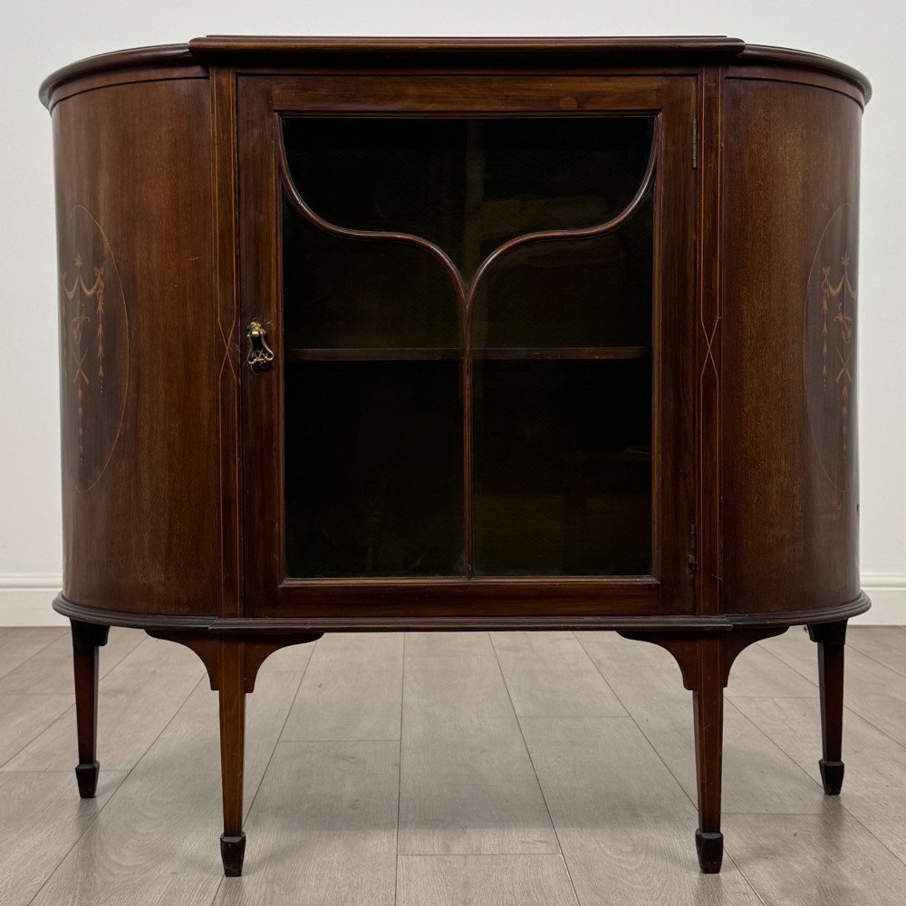 An Edwardian mahogany display cabinet featuring a central glazed door with curved glazing bars, flanked by curved side panels with inlaid floral motifs, raised on tapered legs.