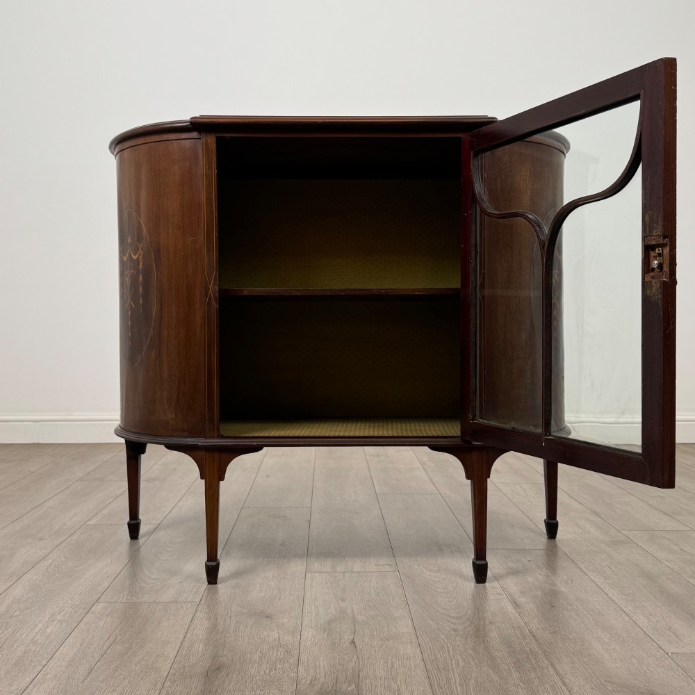 An Edwardian mahogany display cabinet featuring a central glazed door with curved glazing bars, flanked by curved side panels with inlaid floral motifs, raised on tapered legs. View II