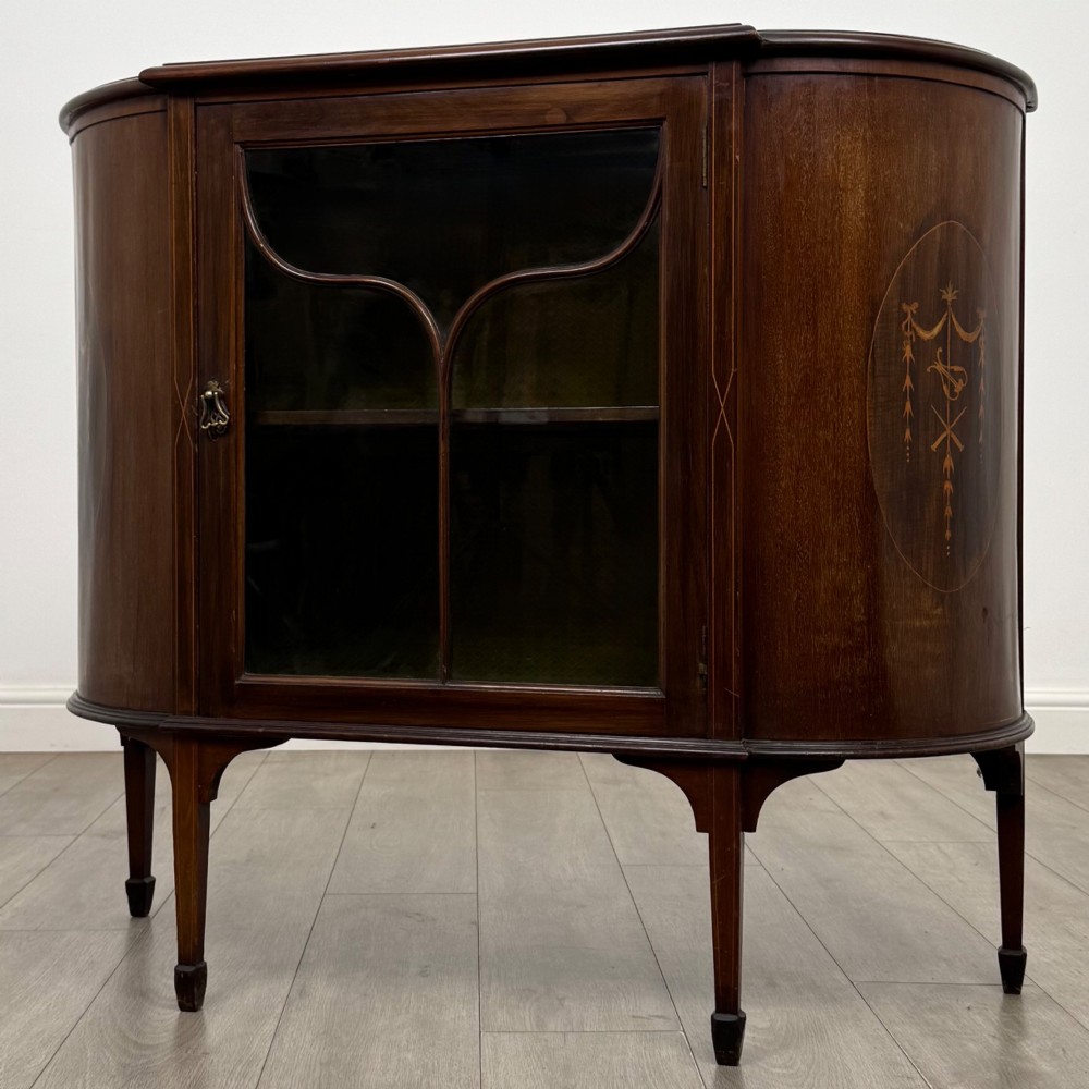 An Edwardian mahogany display cabinet featuring a central glazed door with curved glazing bars, flanked by curved side panels with inlaid floral motifs, raised on tapered legs. View III