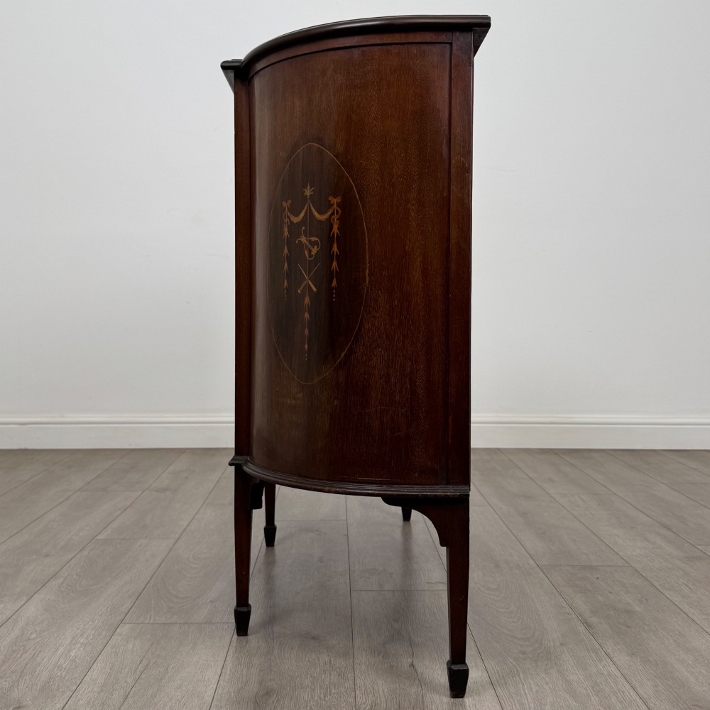 An Edwardian mahogany display cabinet featuring a central glazed door with curved glazing bars, flanked by curved side panels with inlaid floral motifs, raised on tapered legs. View IV