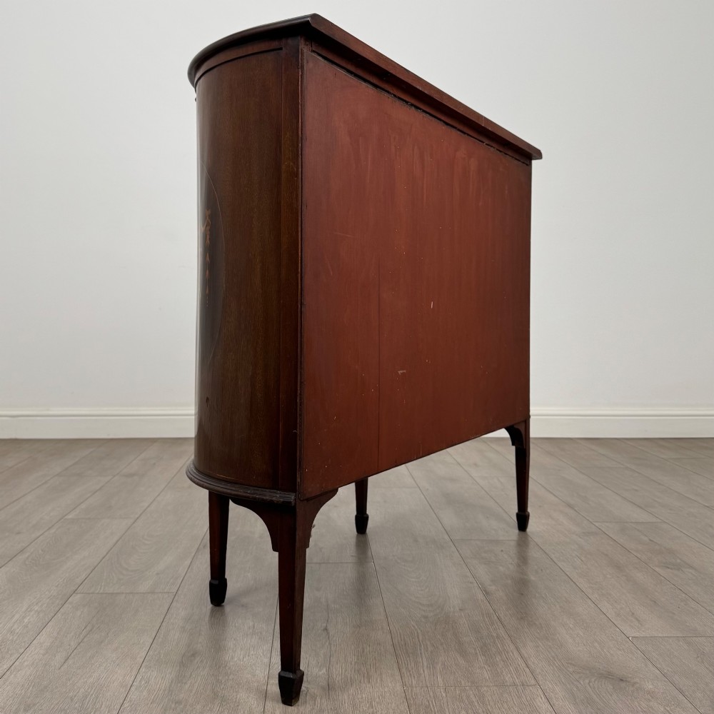 An Edwardian mahogany display cabinet featuring a central glazed door with curved glazing bars, flanked by curved side panels with inlaid floral motifs, raised on tapered legs. View V