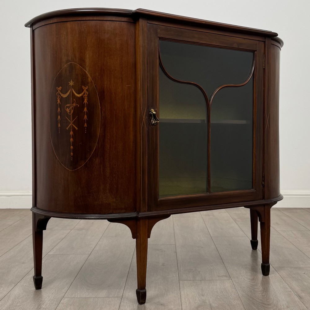 An Edwardian mahogany display cabinet featuring a central glazed door with curved glazing bars, flanked by curved side panels with inlaid floral motifs, raised on tapered legs. View IX
