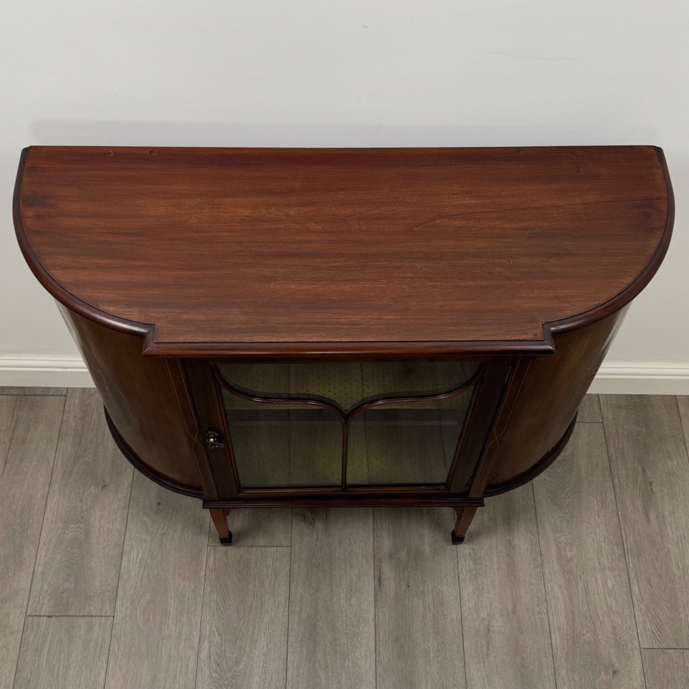 An Edwardian mahogany display cabinet featuring a central glazed door with curved glazing bars, flanked by curved side panels with inlaid floral motifs, raised on tapered legs. View X