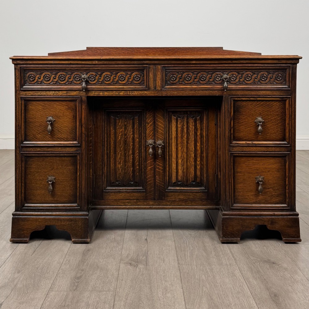 A dark carved oak Edwardian kneehole desk featuring frieze drawers with carved detailing and lower cupboard doors with vertical carved panels.