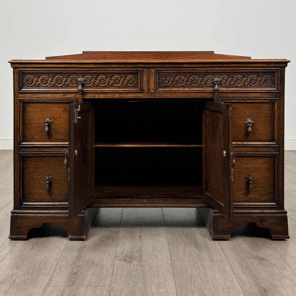 A dark carved oak Edwardian kneehole desk featuring frieze drawers with carved detailing and lower cupboard doors with vertical carved panels. View II