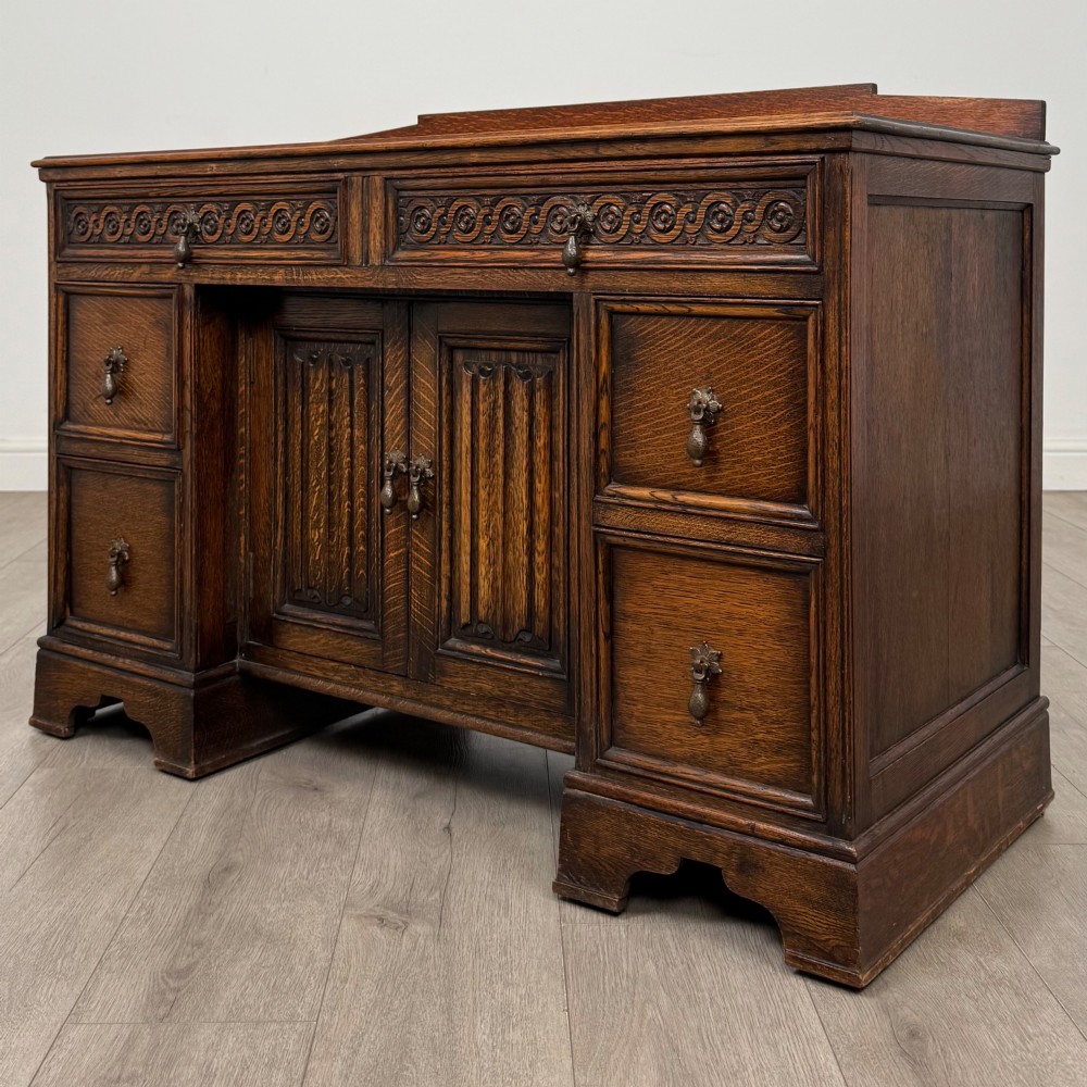 A dark carved oak Edwardian kneehole desk featuring frieze drawers with carved detailing and lower cupboard doors with vertical carved panels. View III