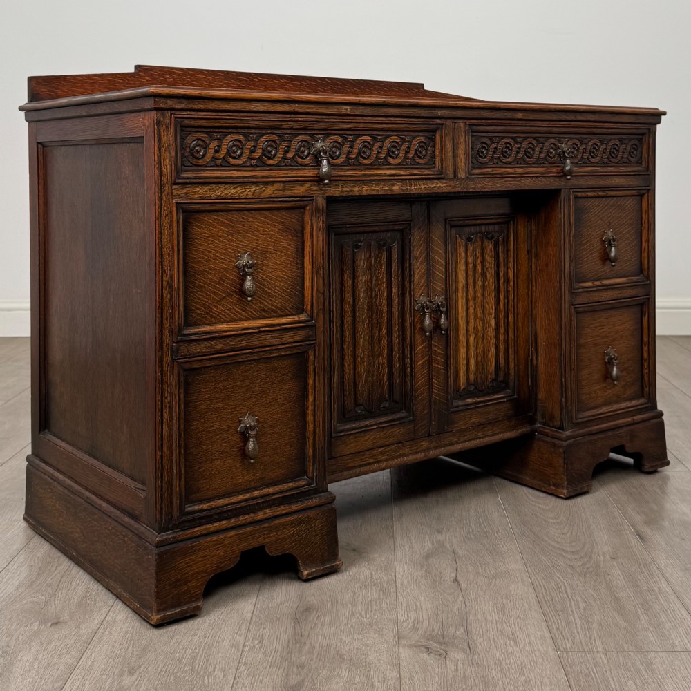 A dark carved oak Edwardian kneehole desk featuring frieze drawers with carved detailing and lower cupboard doors with vertical carved panels. View IX