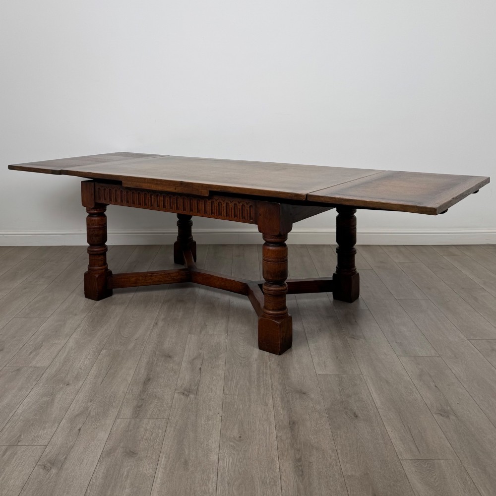 An Edwardian oak draw leaf dining table featuring extended end leaves, carved frieze aprons, heavy turned legs, and an X-form stretcher.