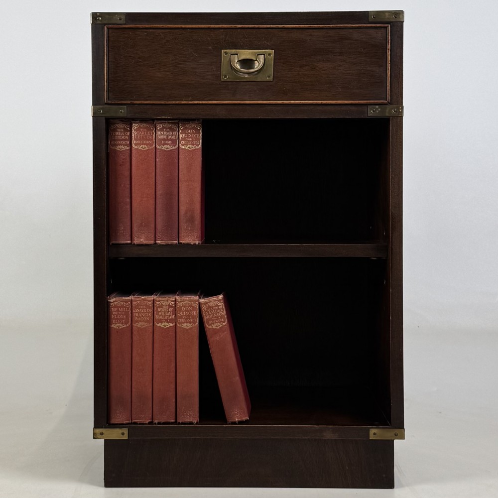 A dark mahogany campaign style cabinet featuring a single upper drawer with a flush brass handle, brass corner mounts, and two open lower shelves.